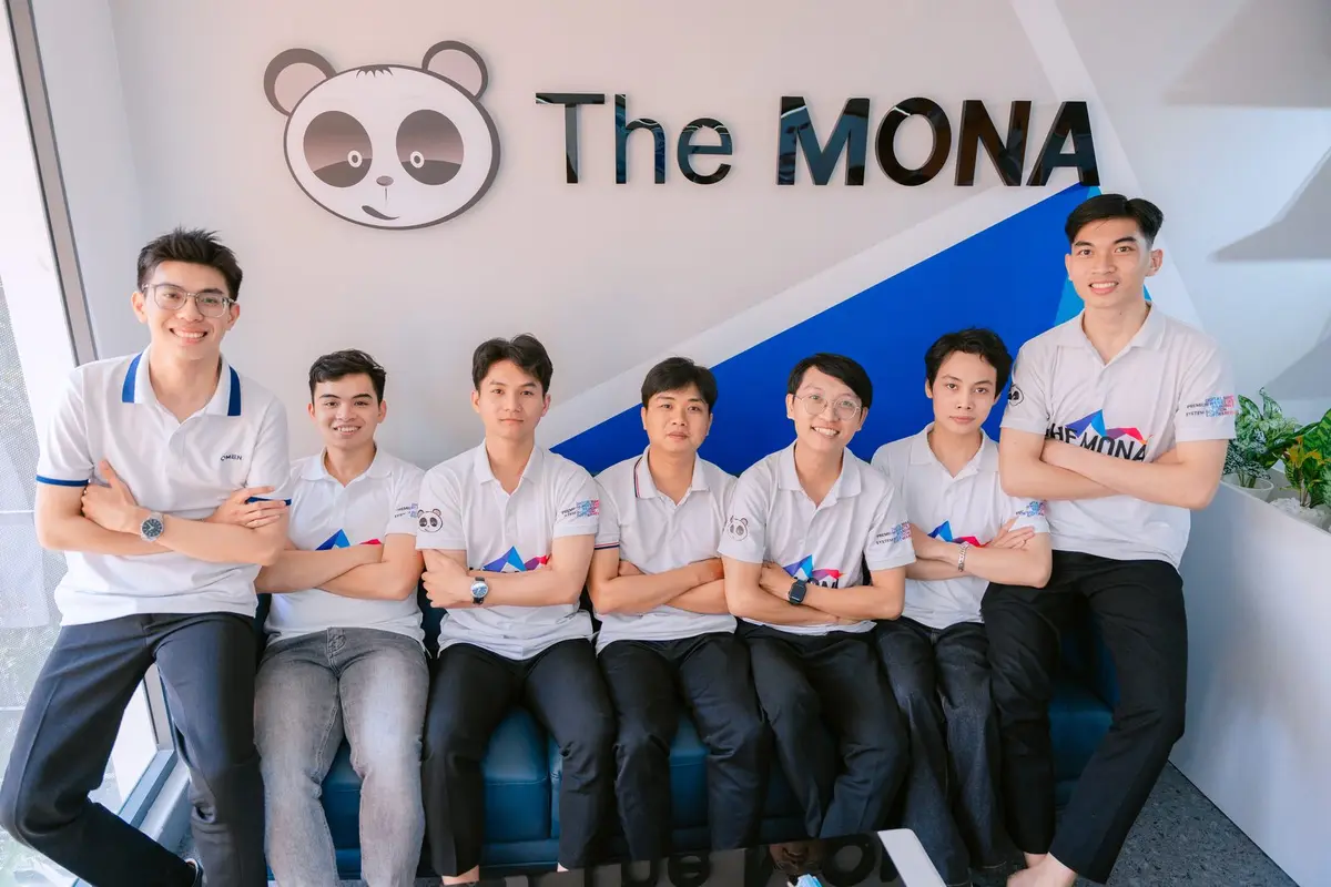 Đội lập trình viên MONA mặc áo THE MONA ngồi dưới logo The MONA ở văn phòng